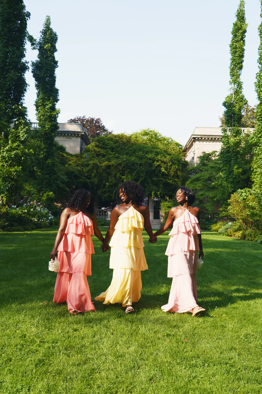 Three women in tiered ruffle gowns in coral, butter yellow and blush, walking hand in hand across a garden lawn
