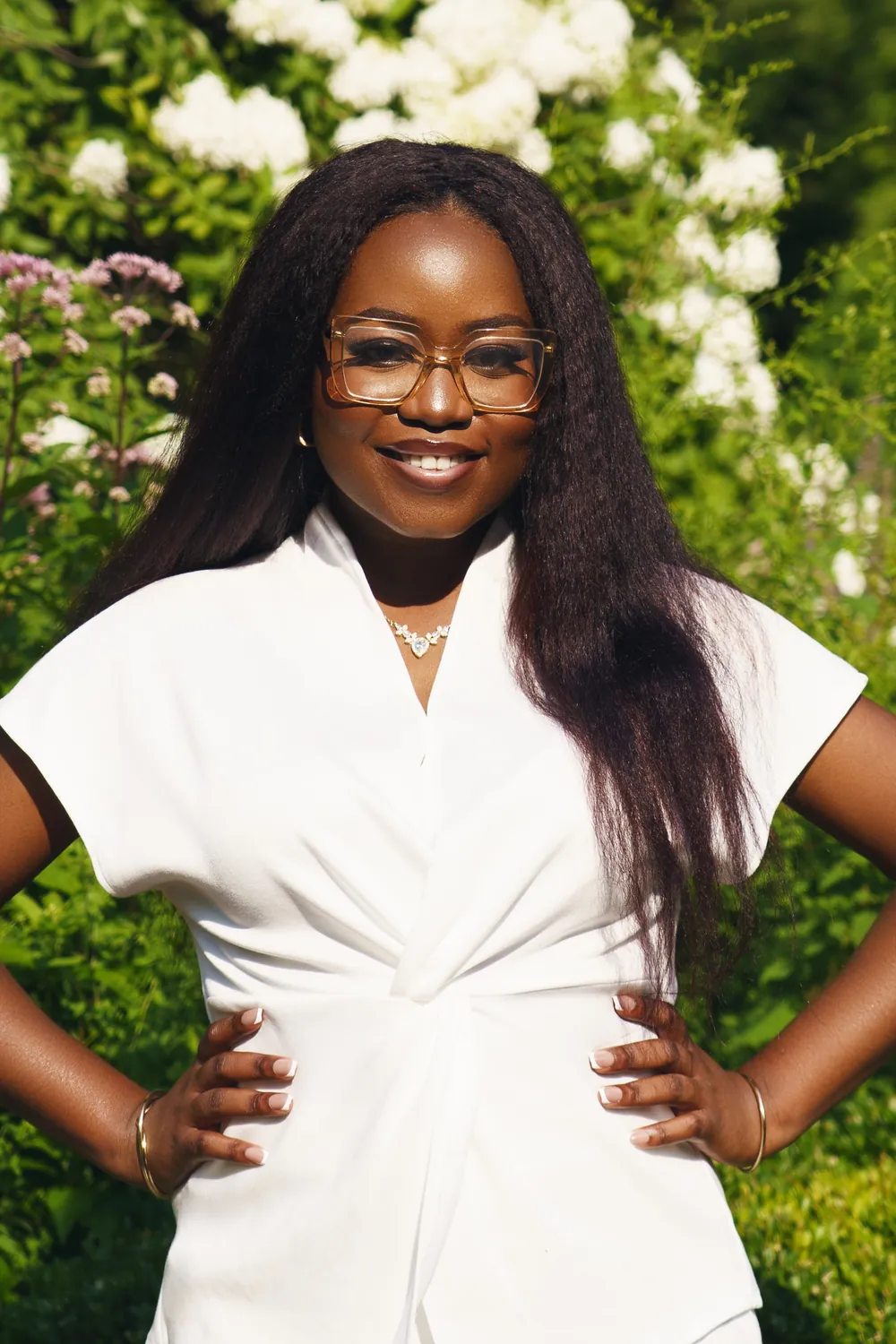 Gemima smiling in a white wrap top, hands on her hips, in front of white hydrangeas